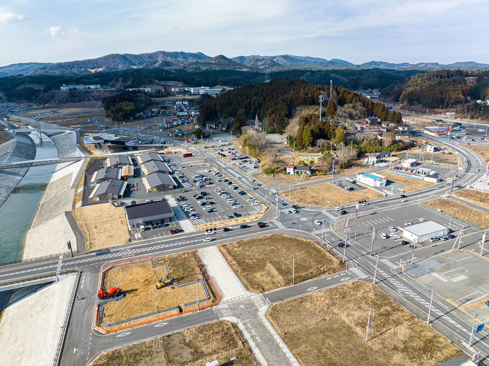 川の右岸に沿って整備された道路や駐車場のある建物群があり、手前には橋が川を横断しており、周囲には舗装された広い道が碁盤目状に伸びている復興地域の昼間の風景を高台から定点カメラで撮影した写真