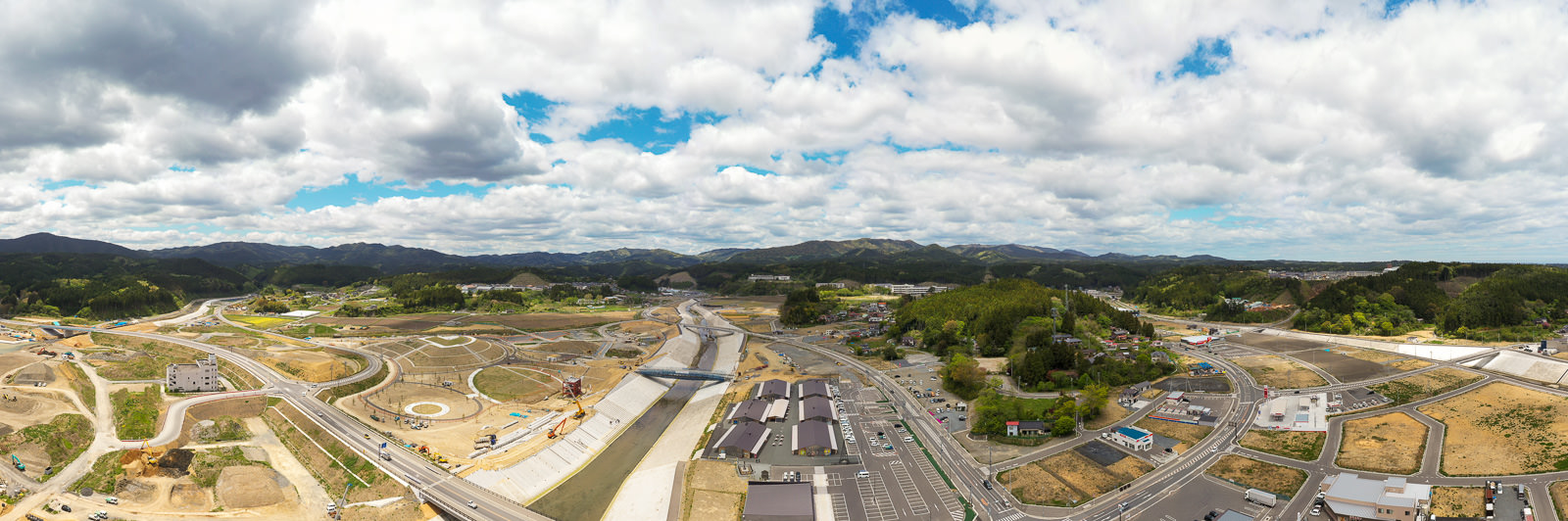 広い青空に雲が浮かぶ下で、山々に囲まれた地域に整備中の土地と道路が広がり、中央には川が流れている様子を写した開発中の山間地域の写真