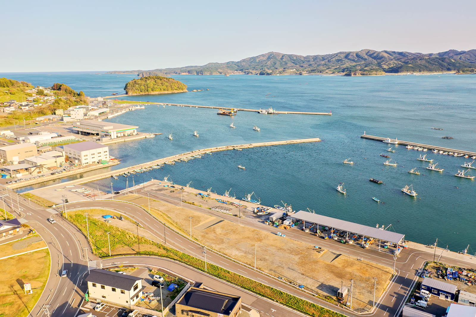穏やかな海に面した小さな港町の風景で、手前には整備された道路と数軒の建物、中央には漁船が停泊する漁港、奥には小島と山並みが広がっており、晴天の下で全体が明るく撮影されている港町の写真