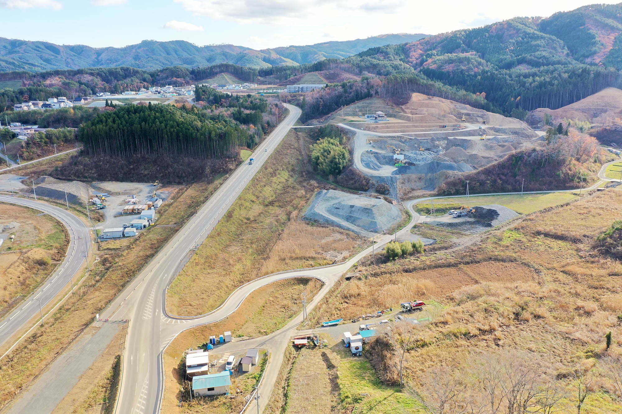 山に囲まれた斜面で盛土工事が進む現場と、それに続く道路、谷あいにある町と山の風景が見える写真