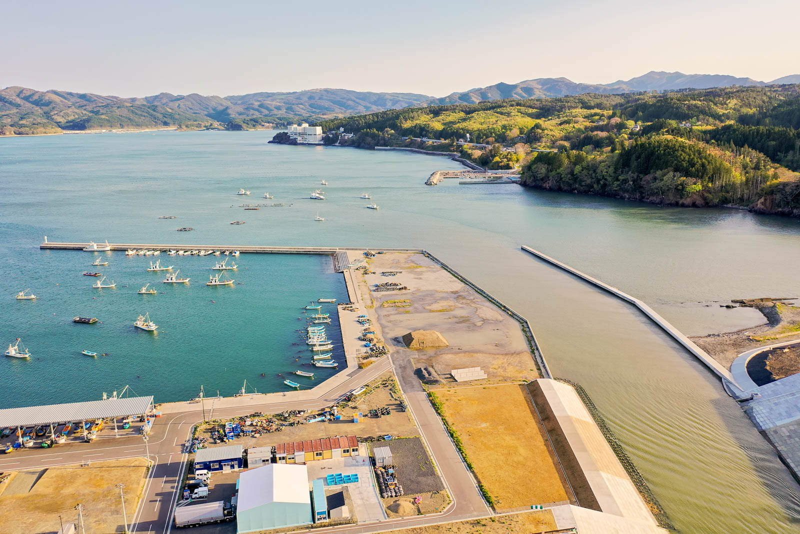 手前に整備された港と建物、中央に漁船が多く停泊する広い湾、右側に川が流れ込む水路と緑豊かな山林、奥には遠くの山々と白い建物が見える風景を、高所から明るい昼間に撮影した港町の写真