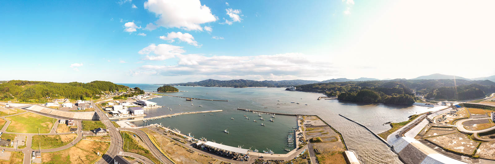 高台から海に面した港町全体を一望し、複数の防波堤と漁船、山並み、湾の奥に続く海岸線が広がる様子を捉えた、夕方の空が美しい湾の写真