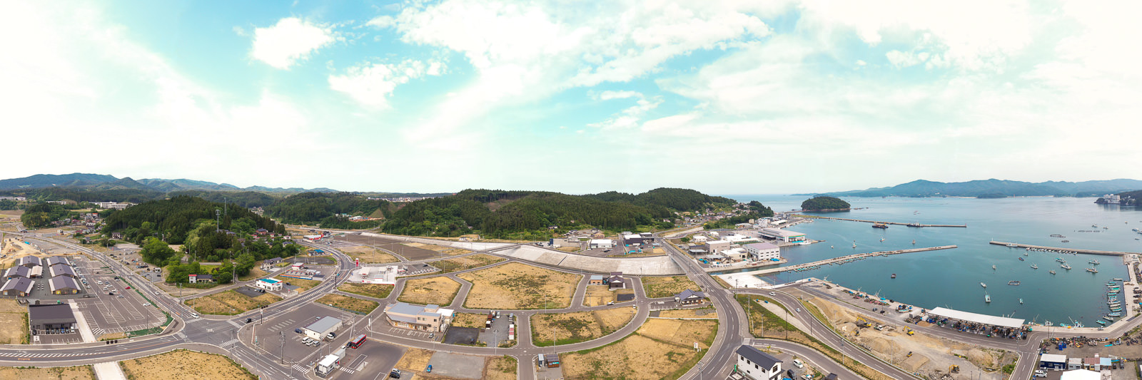 緑に覆われた丘を背景に、建物や道路が広がる港町を俯瞰し、右側に海と停泊中の複数の船が見え、空には青空と雲が混在する写真