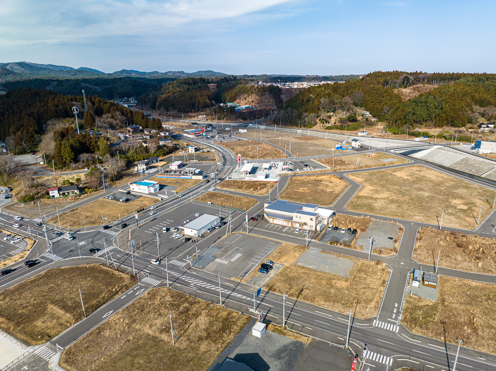 交差点の周辺に商業施設やコンビニが並んでおり、道路は新しく、まっすぐで、空き地も多く見られ、再開発途上であることがわかる復興途中の市街地の昼間の風景を高台から定点カメラで撮影した写真