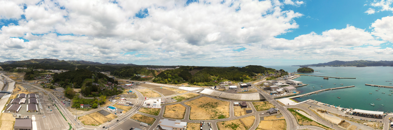 海に面した町を広く見渡すパノラマ写真で、右側には港があり複数の船が停泊し、町には建物や道路、緑地が広がり、背景には丘や山が見え、空には雲が浮かぶ海辺の町の写真