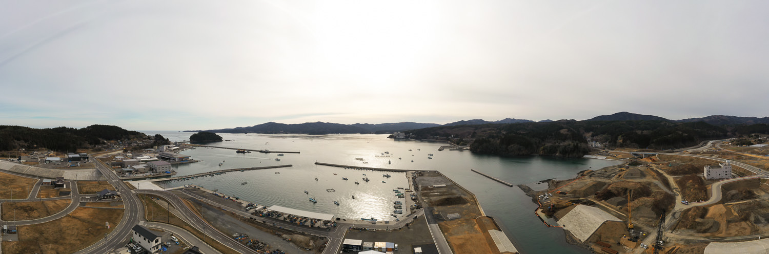 曇り空の下、港に複数の船が浮かぶ海沿いの地域を広く捉えた風景で、周囲には道路や建物、工事現場が点在し、背景には丘や山々が広がっている港湾地域の写真