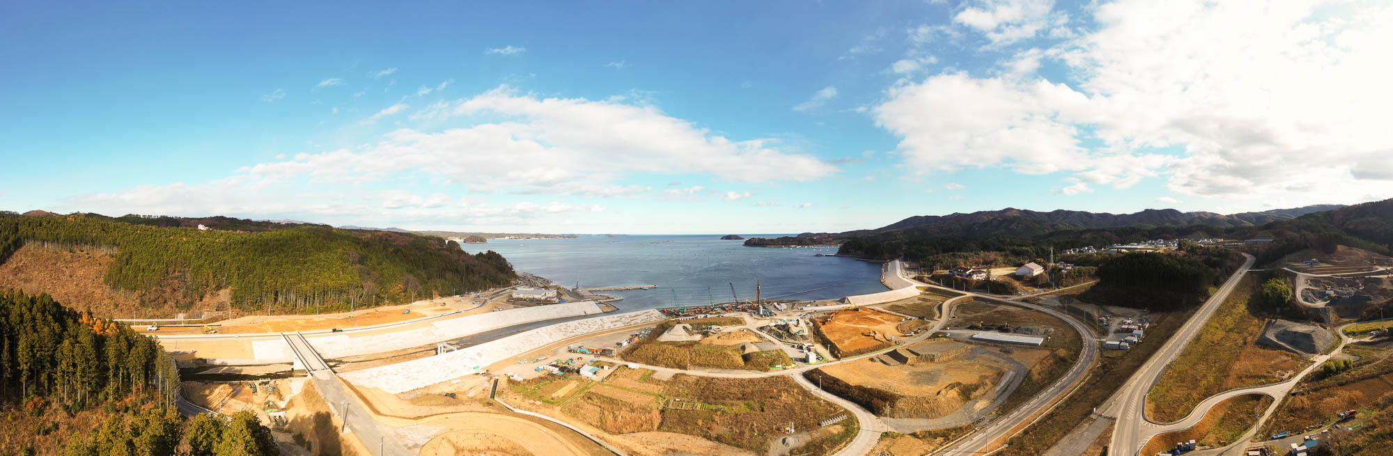 左に山、右に海が広がり、防潮堤が湾を囲むように続き、中央には復興工事が進む土地が広がっている写真