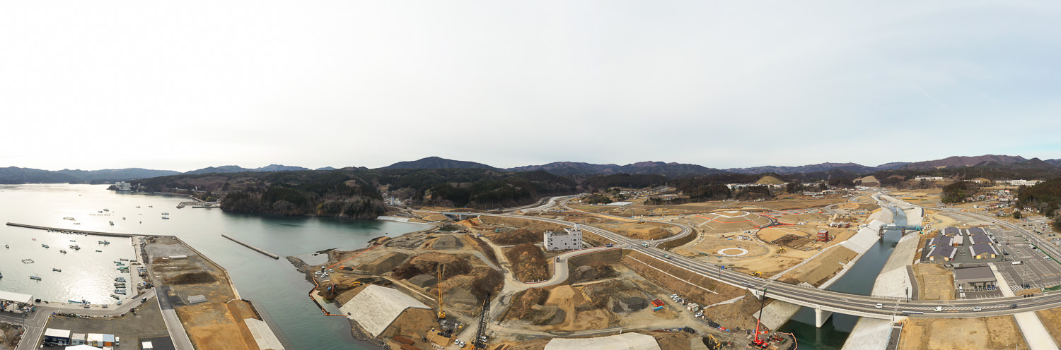 開発または再整備が進む海沿いの地域を広く捉えた風景で、複数の工事現場や道路、インフラ設備が見られ、海岸には船が停泊しており、背景には丘や山々が広がっている沿岸地域の写真