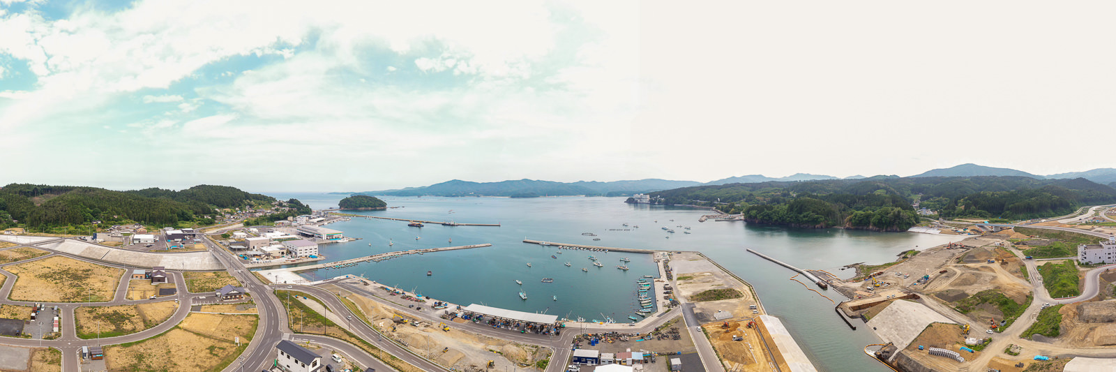 港に複数の船が停泊し、岸辺には建物や構造物が並び、背景に緑の丘と山々が広がり、空が澄んでいる沿岸地域の写真