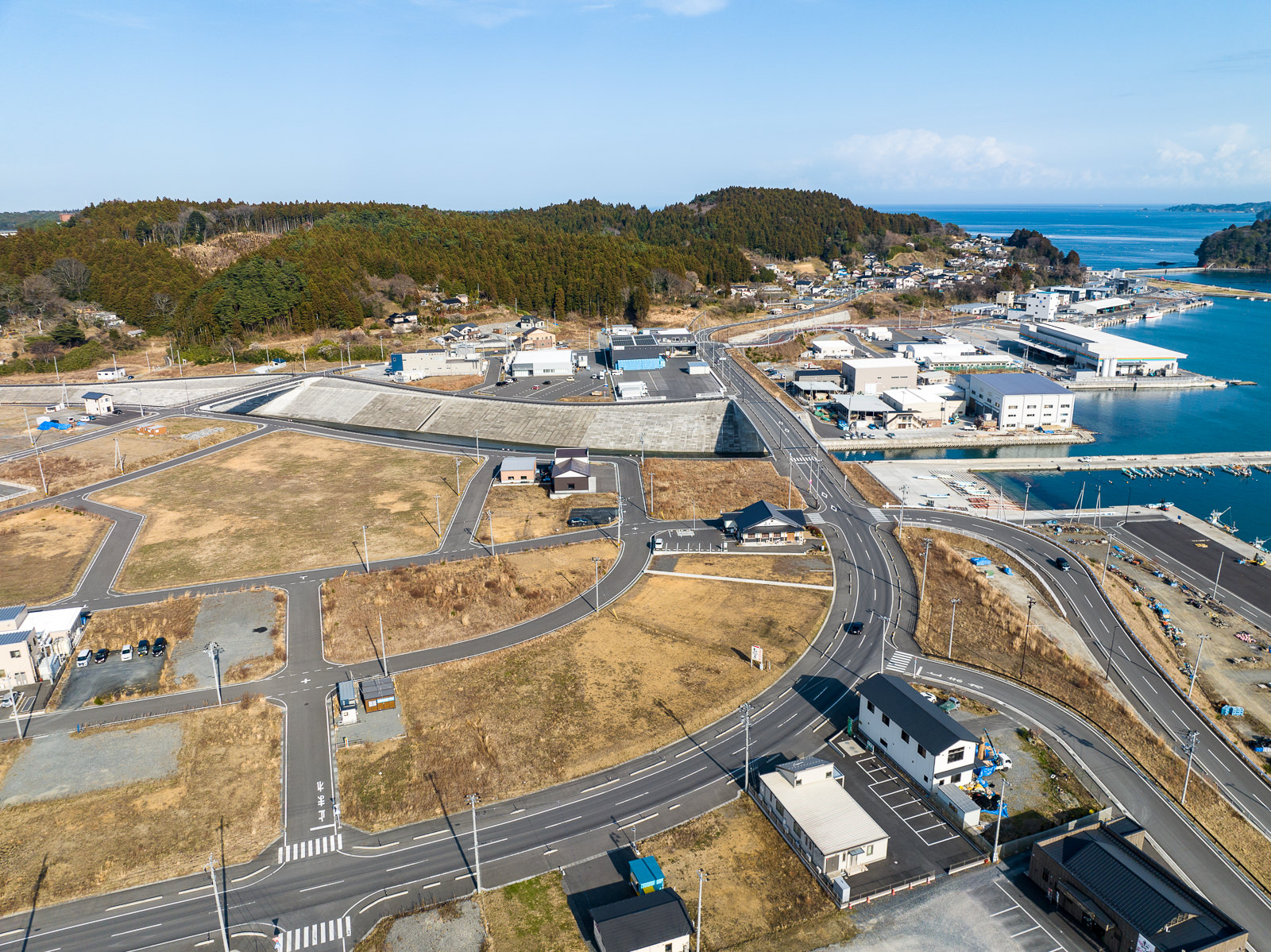 港へ続くカーブを描く道路の周囲に数軒の建物と整備された区画が並び、背景には港の建物群や海が見え、漁港へ続く道と再開発エリアの境目が見える、港近くの市街地と海への接続部の昼間の風景を高台から定点カメラで撮影した写真