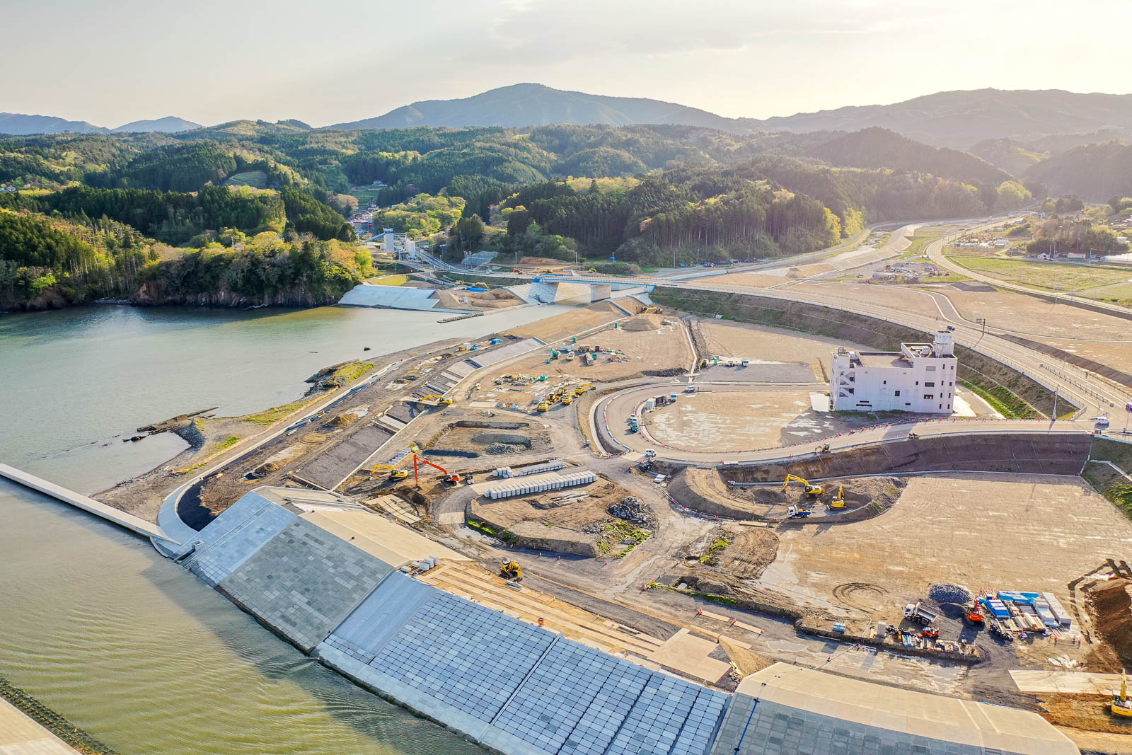 手前に川沿いの護岸工事が進む広大な造成地が広がり、中央には重機が多数稼働する建設現場と白い防災庁舎が立ち、奥には緑豊かな山々と蛇行する道路が続く様子を夕方の柔らかな光の中で撮影した復興工事中の町の写真