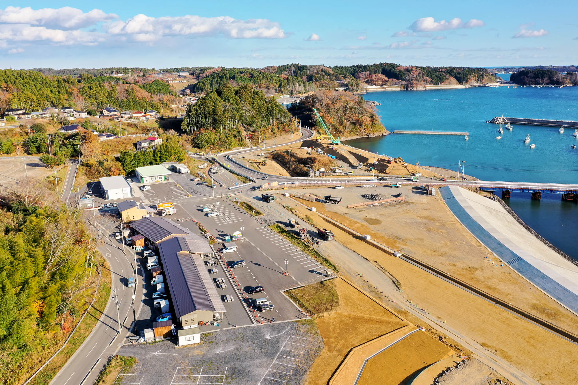 左下に駐車場と複数の建物があり、中央には整備中の道路や護岸工事が進む様子があり、奥には青い海と港湾施設が見える写真