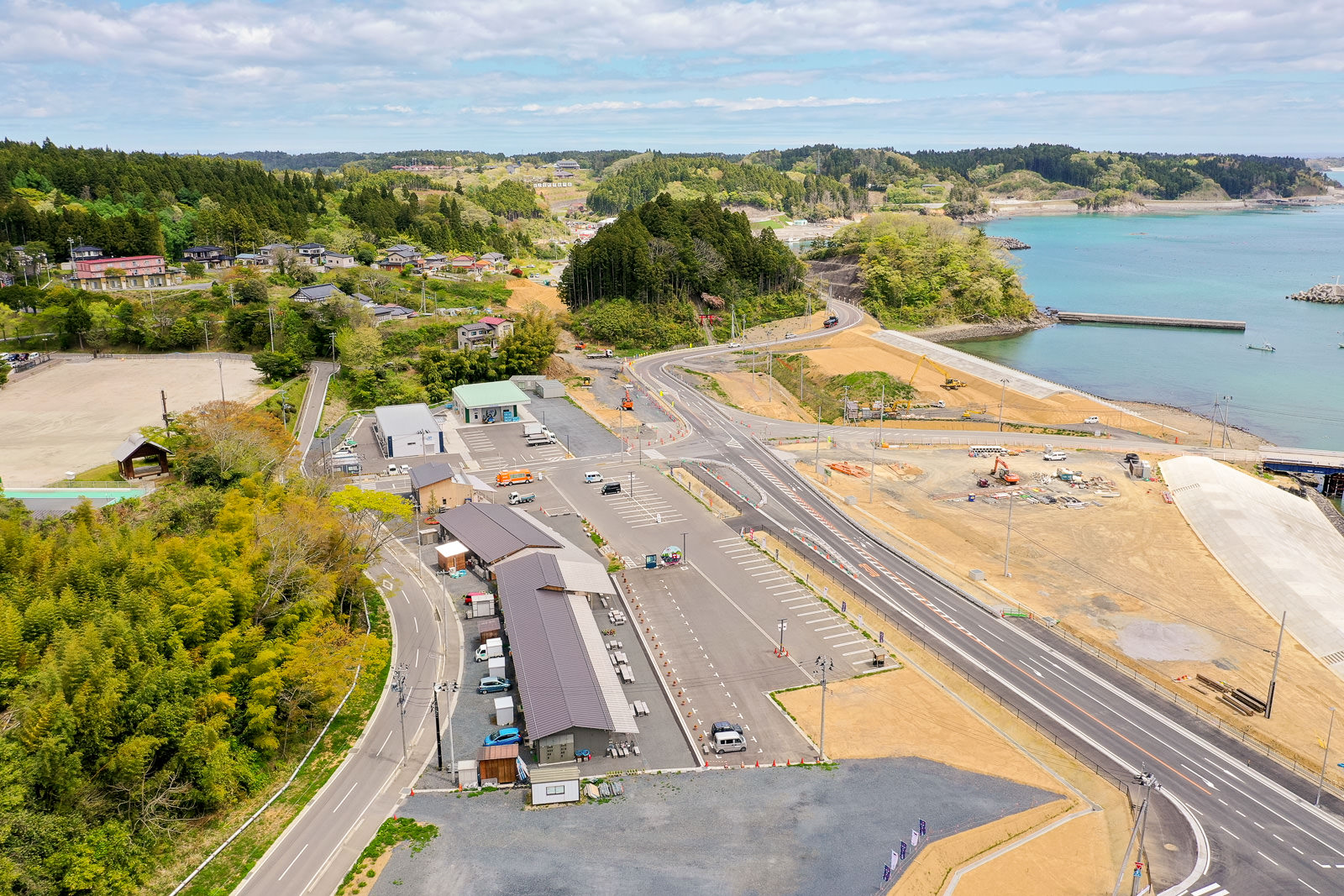 海沿いに道路と駐車場が広がり、周囲には緑豊かな山々と点在する建物が見える風景の写真