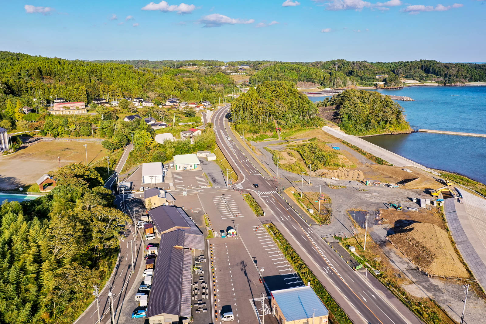 左下に駐車場と建物群、中央から右奥にかけて湾へ向かう道路と工事中の土地が広がり、奥に青い海と緑の山が見える港に向かう道路と工事現場の写真