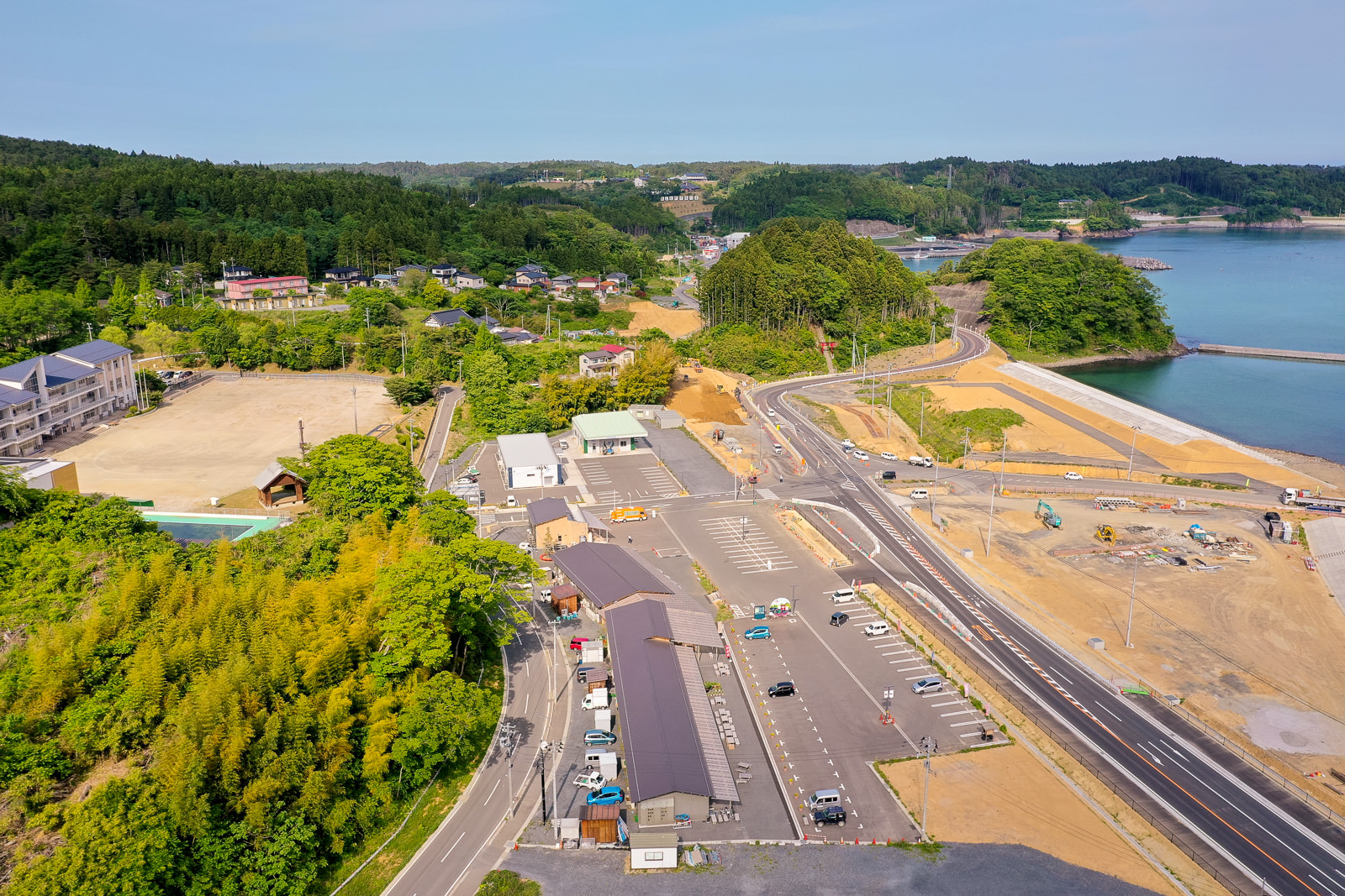 緑豊かな山々と海に囲まれ、駐車場や建物が点在する沿岸地域を広く見渡した写真