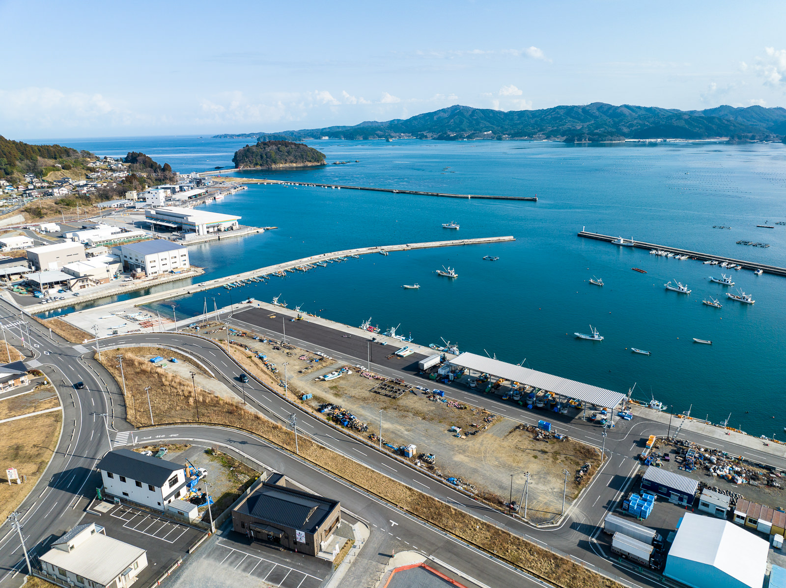 右側の海沿いには漁業施設や倉庫が建ち並び、多くの小型漁船が岸に停泊しており、湾内は波が穏やかで、背景は山と海の境界が広がる漁港と作業施設の昼間の風景を高台から定点カメラで撮影した写真