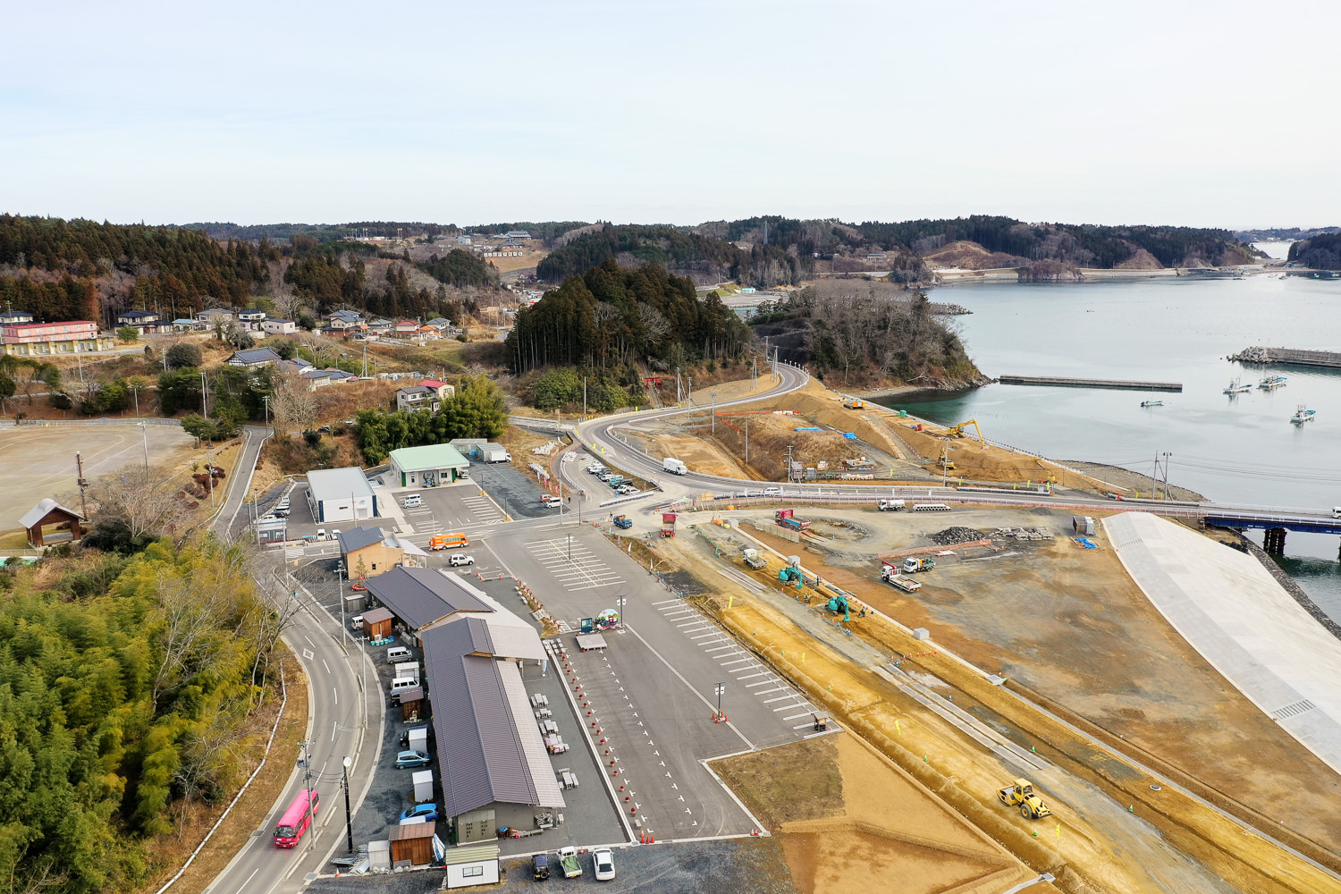 海沿いの町を空から見下ろした写真で、手前には複数の建物や道路、駐車場が広がり、奥には船の浮かぶ水域、木々に覆われた丘陵地、橋や工事現場などのインフラが見える写真