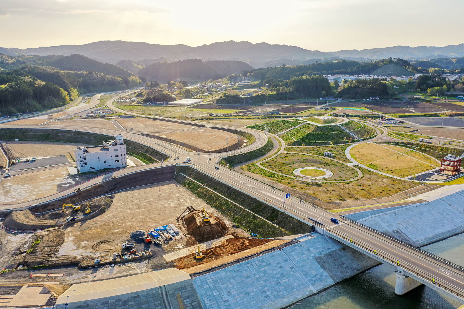 手前に川にかかる大きな橋と護岸工事現場、中央には白い防災庁舎と複数の重機が稼働する造成地、右側には円形の歩道が特徴的な整備された公園、そして奥には山並みと住宅地が広がる風景を夕方の柔らかな光の中で撮影した復興が進む町の写真