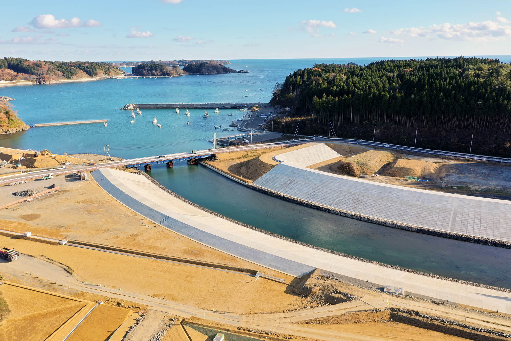 曲がりくねった護岸が海から続く川を囲み、奥には防波堤に囲まれた港と小型船が浮かぶ海が広がり、周囲を森林に囲まれている様子の写真