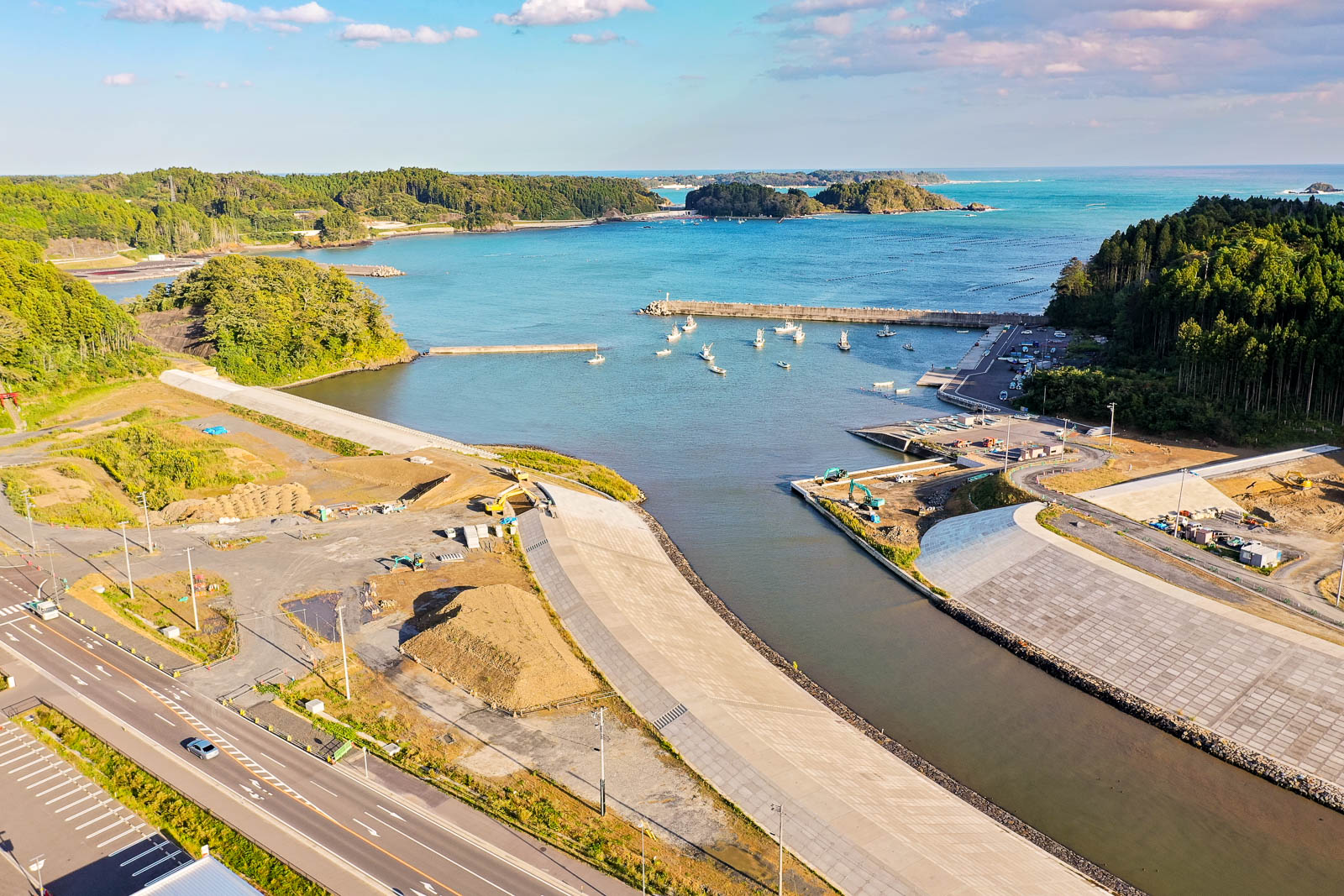 中央に河口と湾、湾内に並ぶ小型漁船、両岸に防波堤と工事中の施設が見える防波堤の整備状況が見える写真