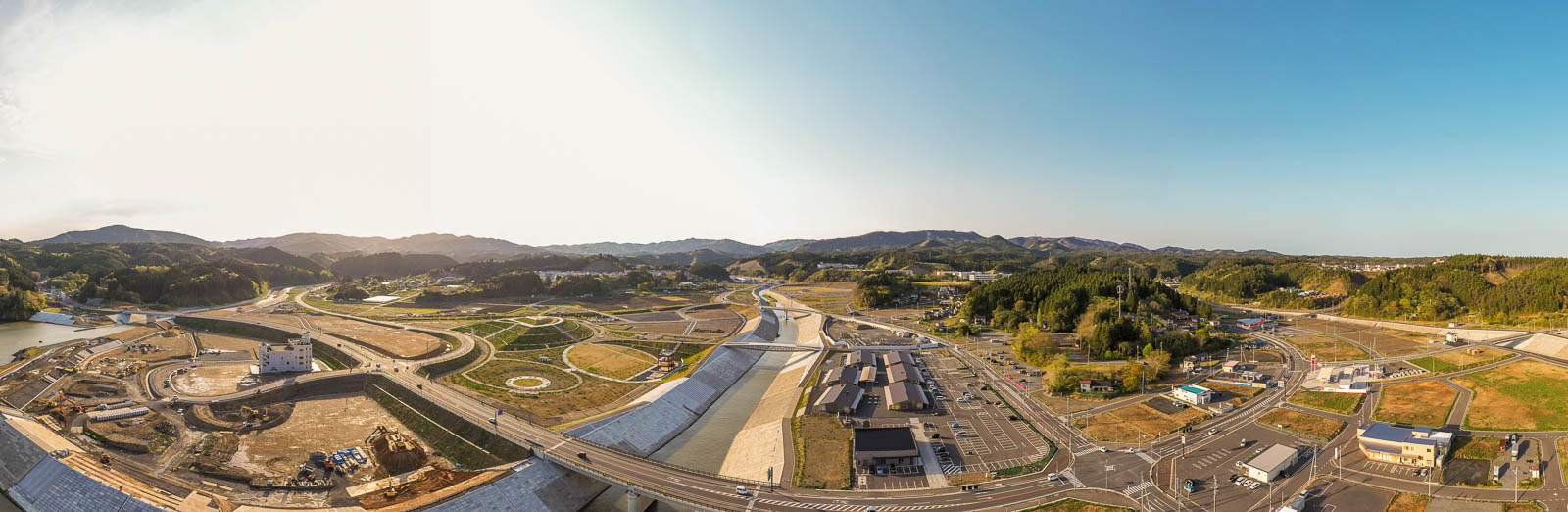 中心に川と堤防、その右側に新しく整備された施設群と道路網が広がっているパノラマ写真