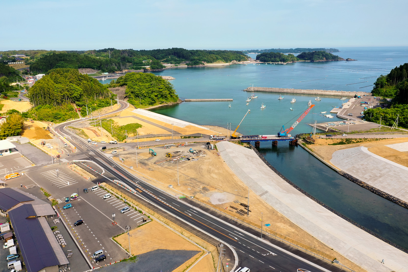 港にクレーンや建設機材が並ぶ沿岸の工事現場を空から見下ろし、道路や駐車場が広がり、周囲には緑の丘と水域があり、港には船が停泊している写真