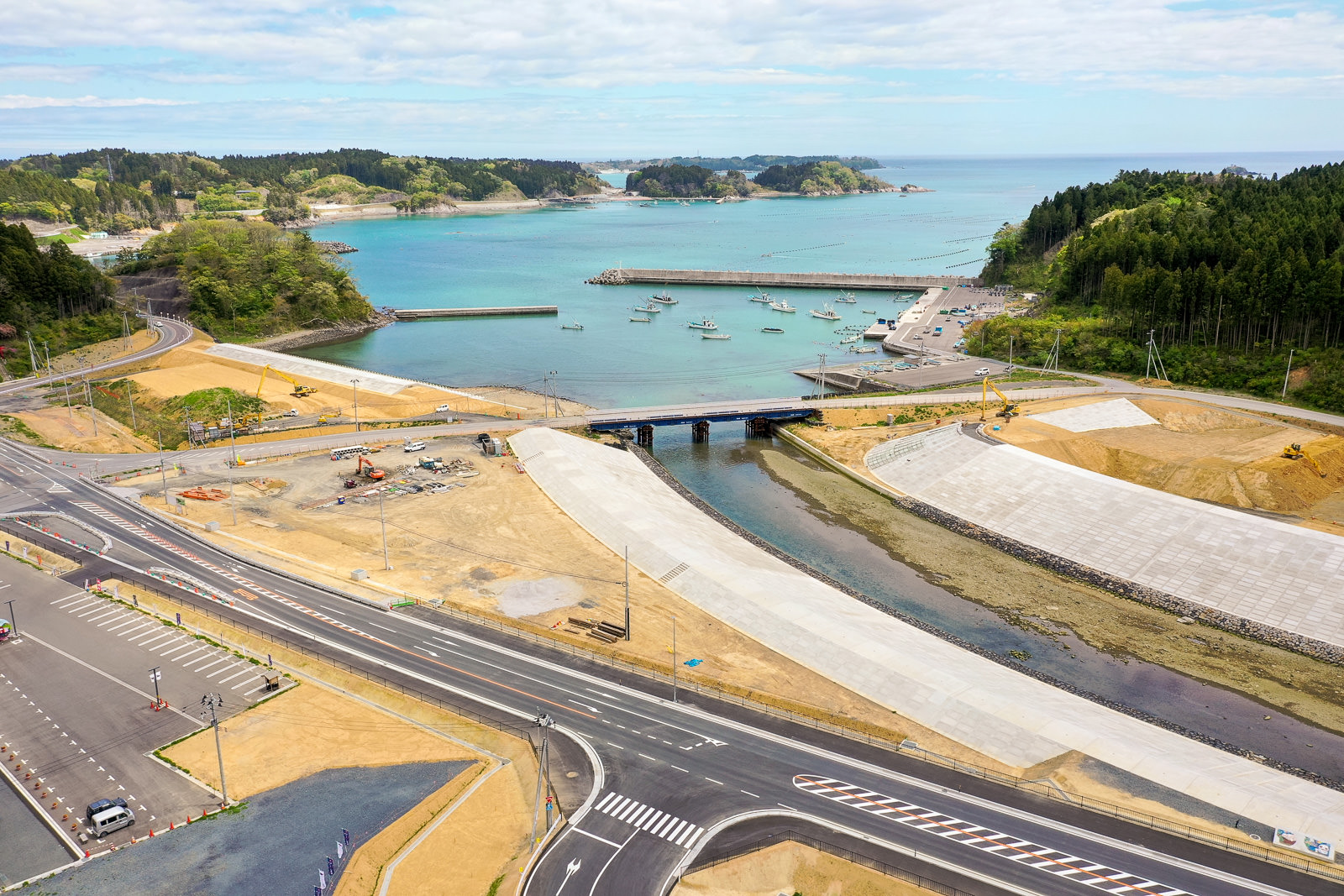 小さな港に複数の船が停泊し、交差点や車線が整備された道路と橋が水路をまたぎ、周囲には緑の丘や工事現場が広がる海辺の町の写真