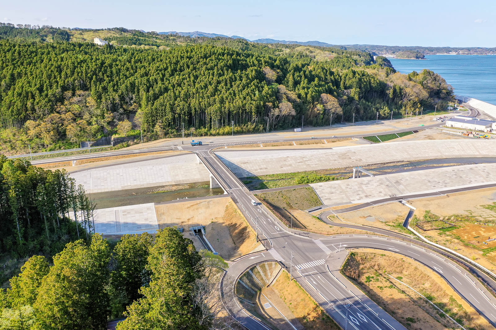 手前には立体的な道路構造があり、緩やかなカーブと交差点、その奥にまっすぐ伸びる橋が見え橋の下にはコンクリートで覆われた川が流れており、左右両側とも高く広い護岸が整備されている写真
