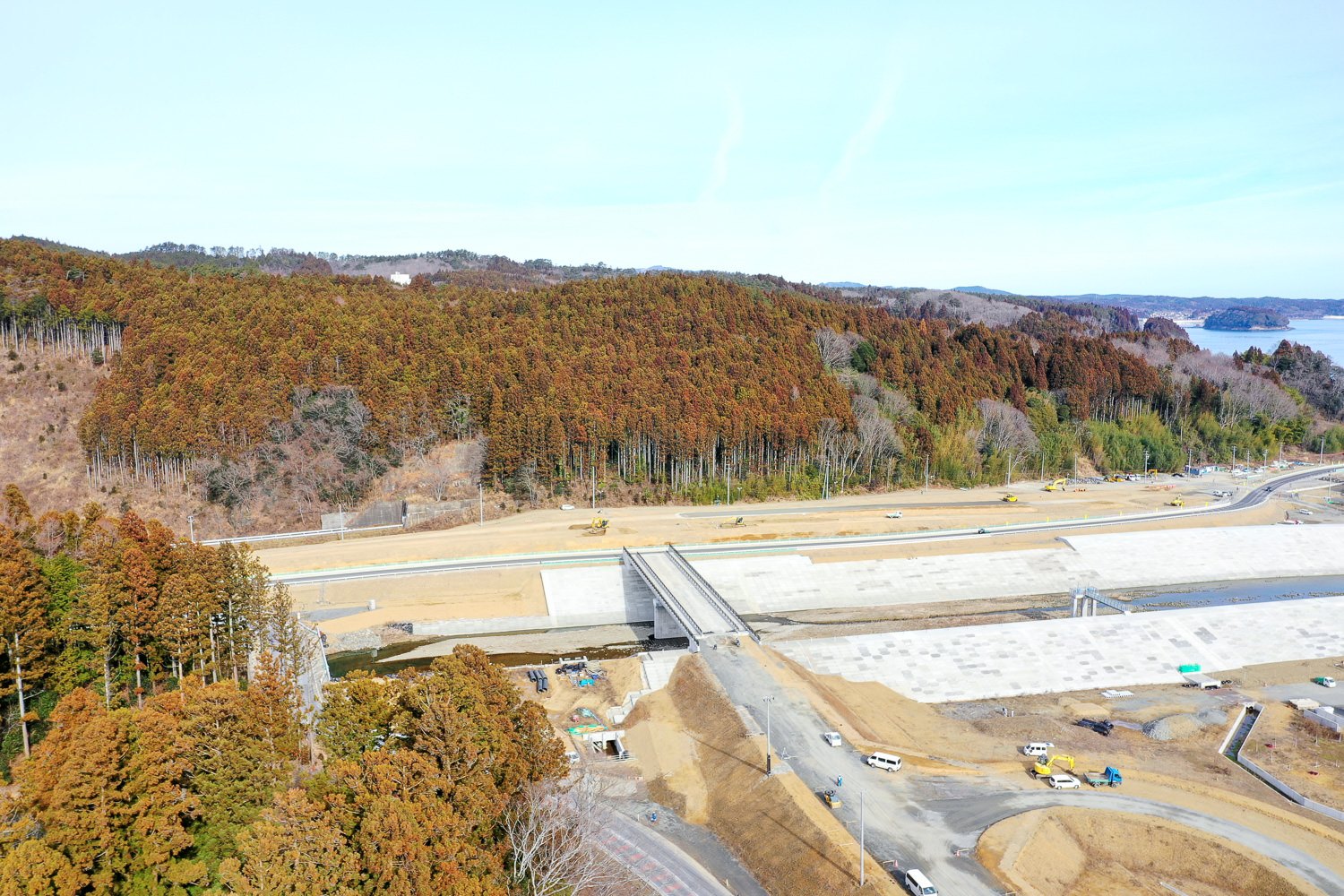 青空の下、道路の上を橋が横切る風景で、橋の両側には紅葉した木々が広がる森林があり、周囲には工事車両や機材が見られる開発中の地域の写真