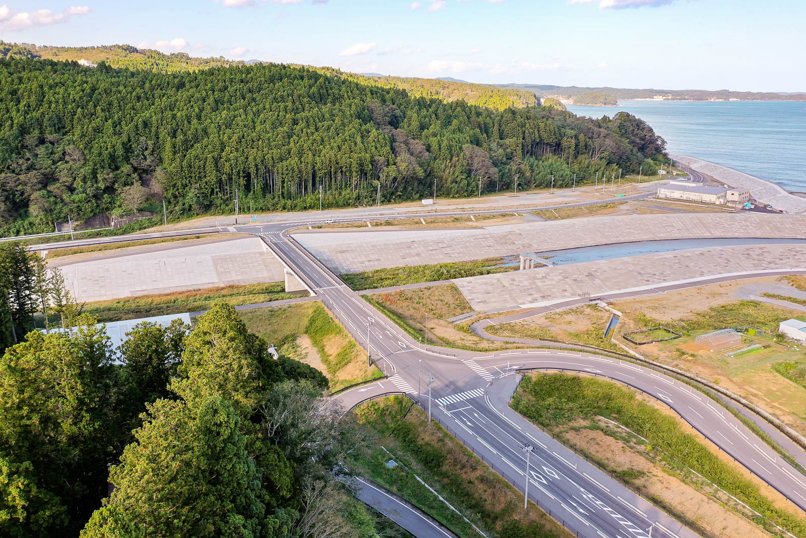 防潮堤に囲まれた地域の中央に小さな川が流れ、複数の橋がその川を横切っており、その両側には山と海が広がる静かな景観が広がっている写真