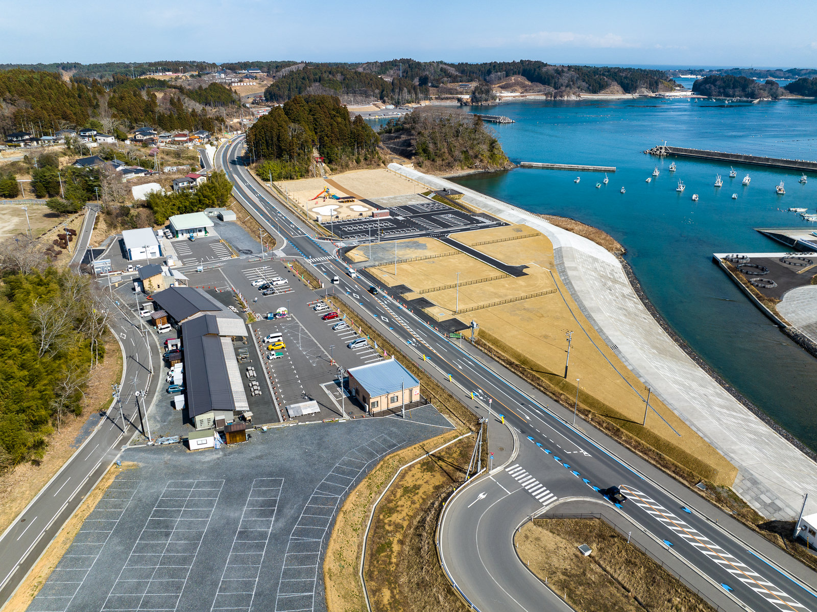 海岸付近に整備された道路と駐車場に囲まれた小規模な建物群が並び、奥に海と防潮堤が見える昼間の風景を高台から定点カメラで撮影した写真