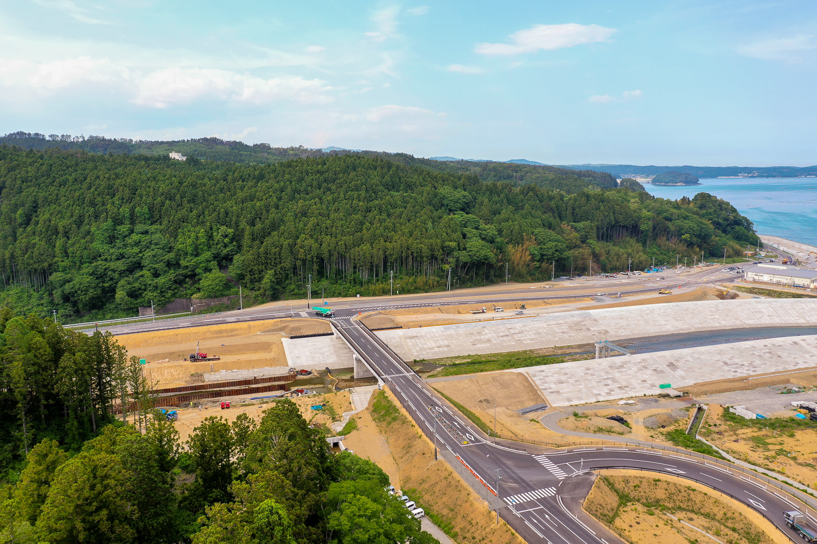 新しく整備された道路の交差点が手前に広がり、周囲には緑豊かな森林の丘が広がり、背景には海岸線が見え、工事車両や機材が点在する開発中の地域を晴天のもとで空から見下ろした写真