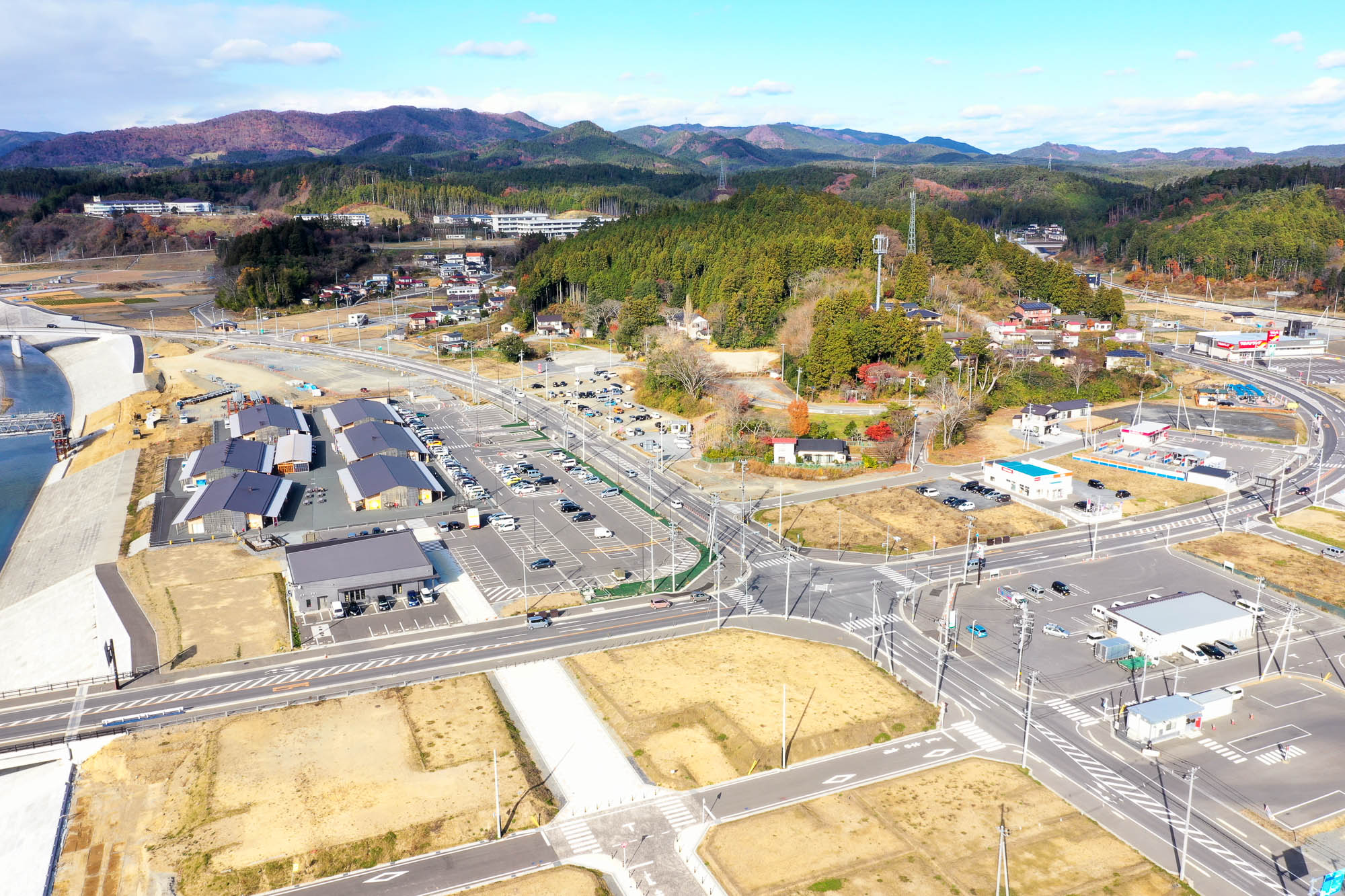 中央の交差点を中心に道路が碁盤の目のように整備され、仮設商店街や駐車場、周囲に点在する建物や紅葉の山々が見える様子を空撮した写真