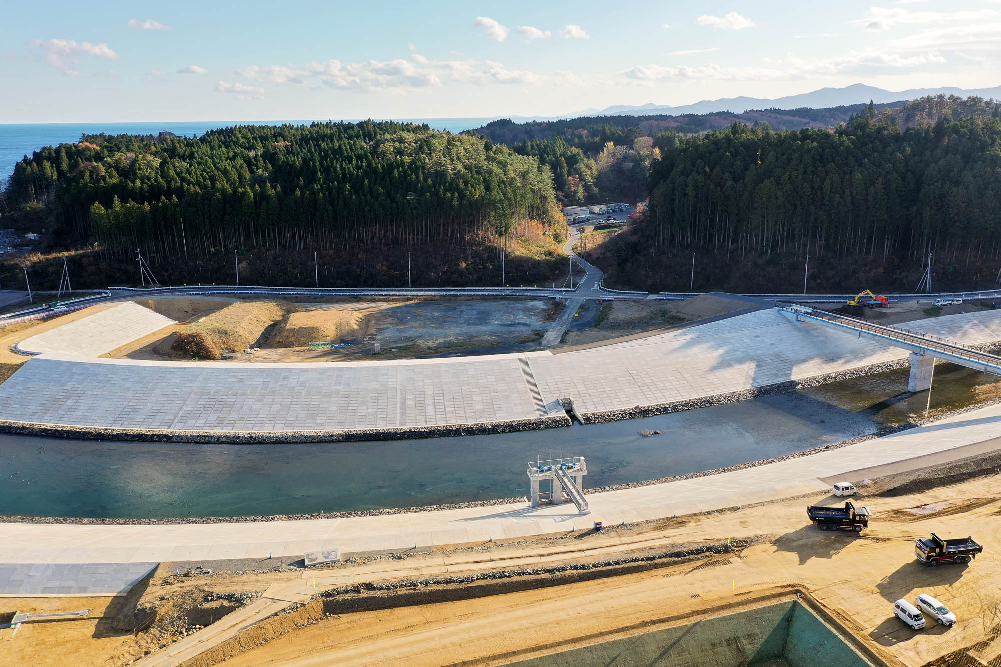 右下に建設車両が並び、中央には川にかかる橋と整備中の護岸が続き、背景に山と森林が広がっている写真
