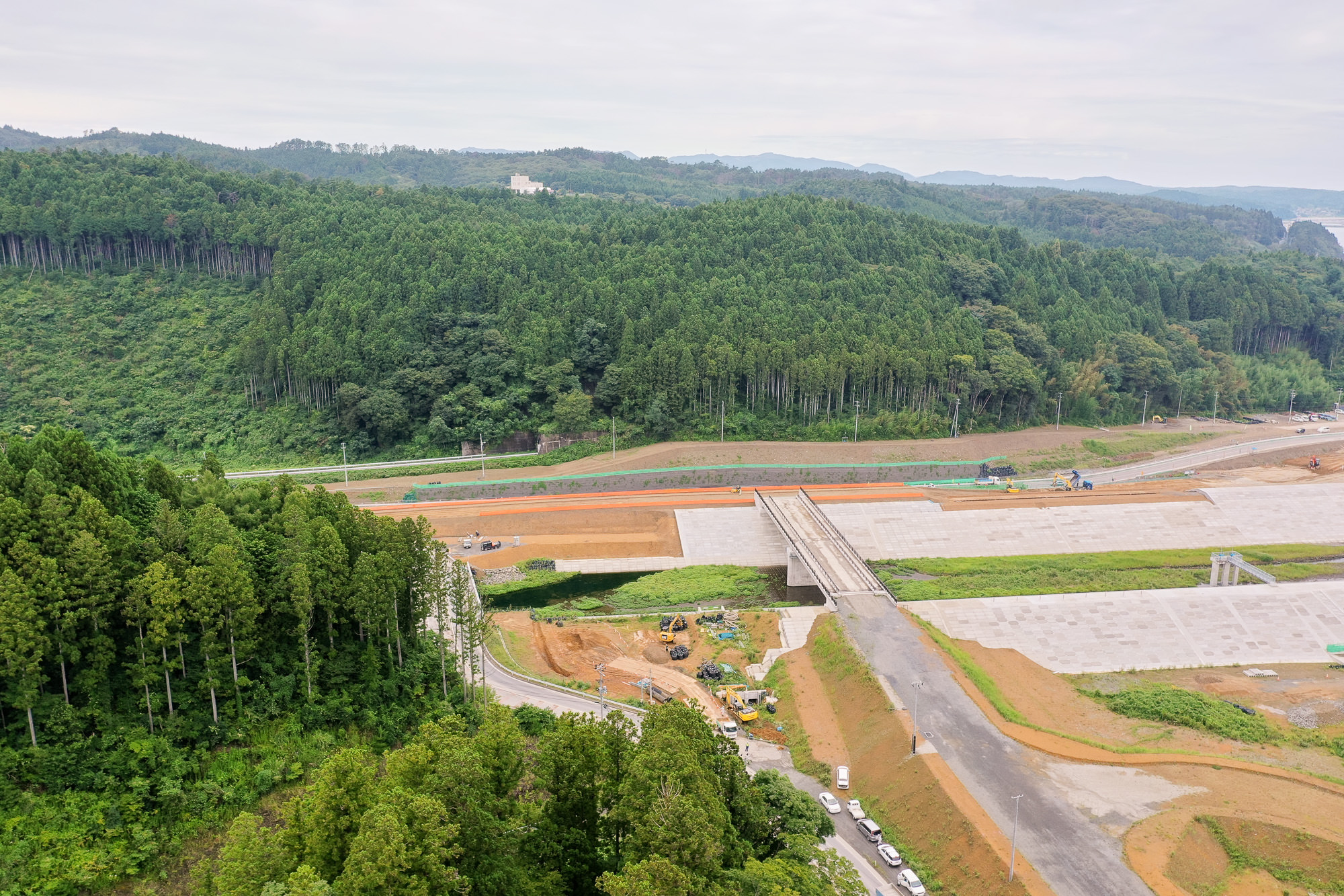 岸辺に並ぶコンクリート護岸区域、背後に緑の野菜畑、遠景に山と海岸線が交互に見える風景