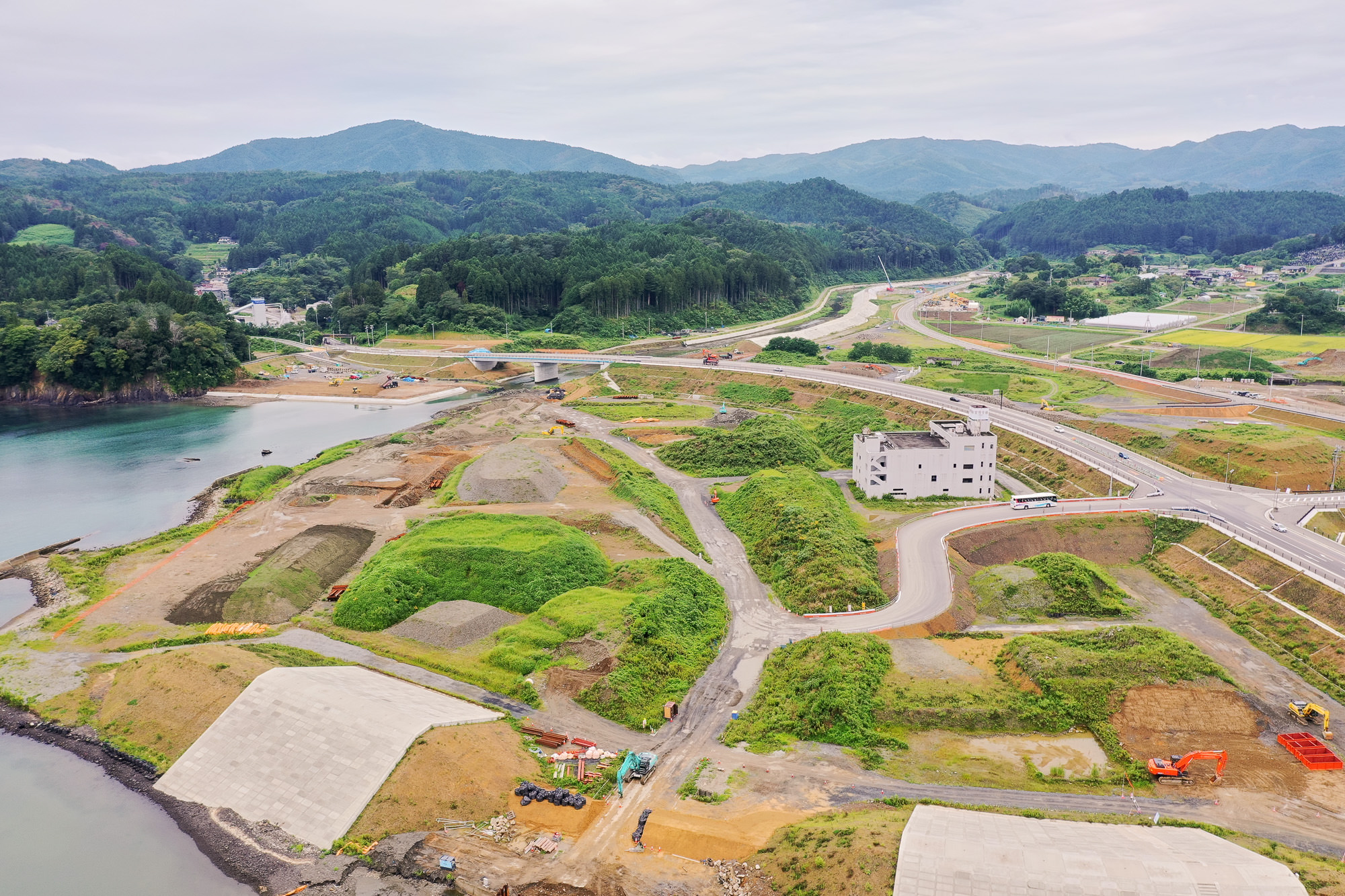 海岸沿いの造成地と曲がりくねる新設道路、中央の白い廃墟のような建物と、背後に連なる山並みや集落が見渡せる開発中の地域の写真