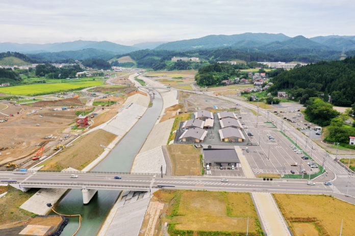 中央にコンクリート護岸の川と橋、右側に工事中の商業施設と駐車場、遠方に森と住宅地、さらに奥に山並みと海が望める構図の写真
