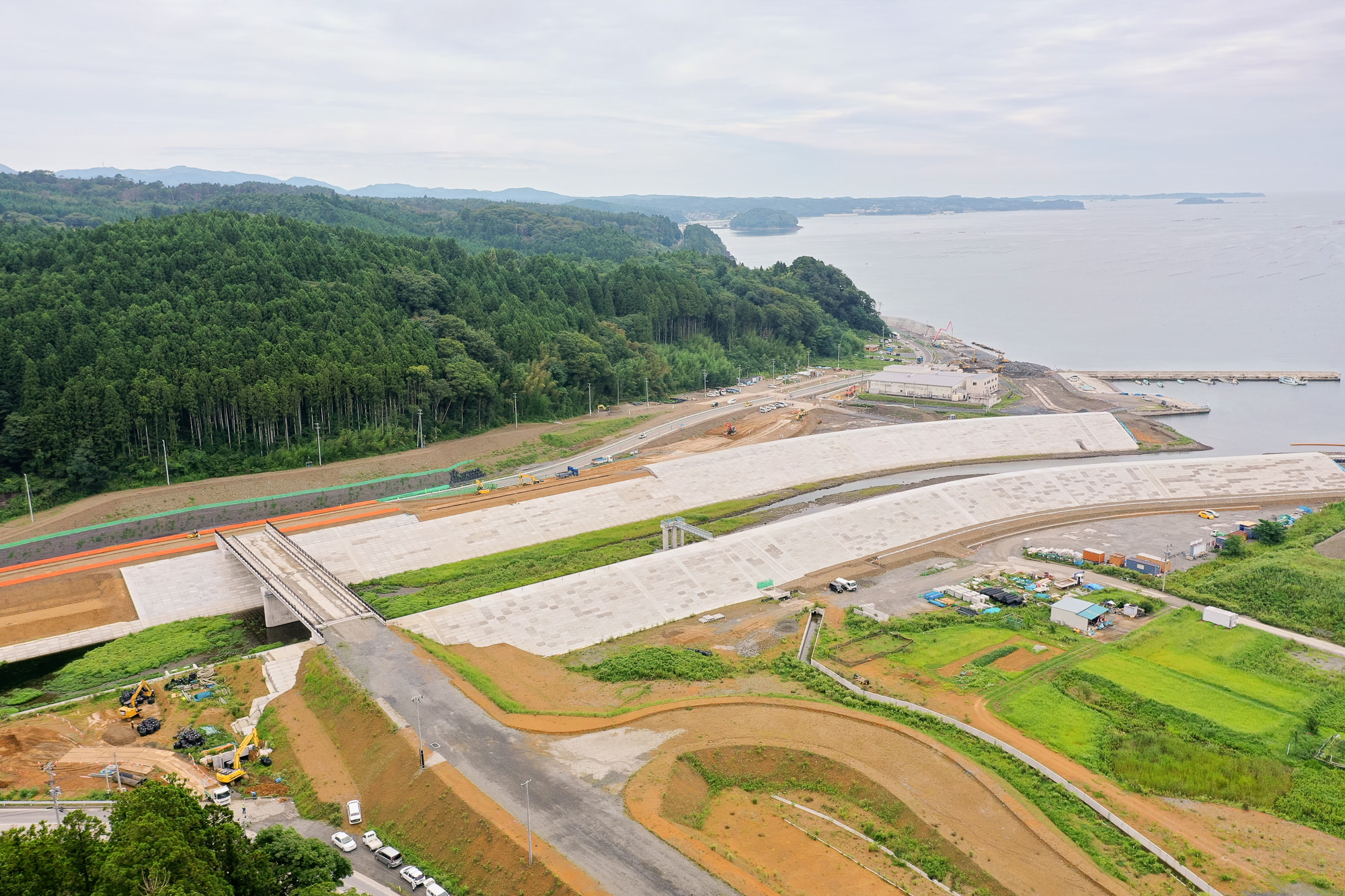 中央に湾に向かう道路網と造成地、遠くに住宅地と丘陵、海と港の設備が写る全景写真