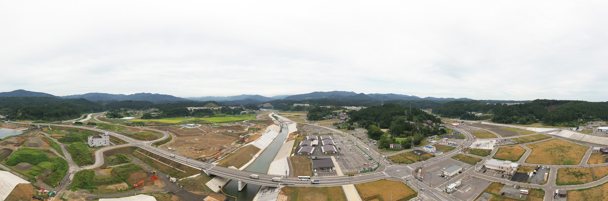 川と交差点がある広い再開発中の平地に道路が通り、奥には田畑や山が広がっている様子を高所から俯瞰した気仙沼市内陸部の写真