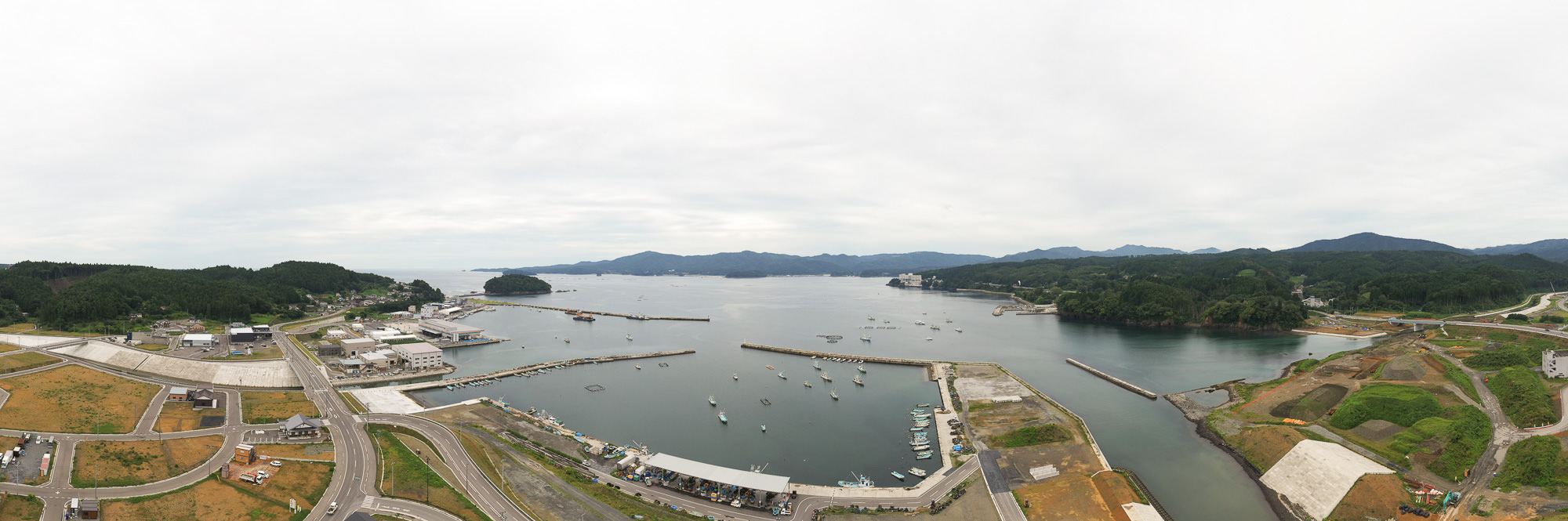 港に沿って広がる街並みと山々に囲まれた湾を広範囲から俯瞰し、湾内には多くの漁船や防波堤が見え、奥には島と対岸の山並みが広がる風景の写真