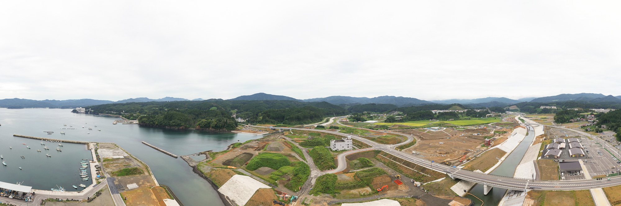 右側の湾と湾に面した林に囲まれた小さな入り江を含む自然豊かな海岸線と、港の背後に広がる整備中の土地と山々が写された沿岸部の写真