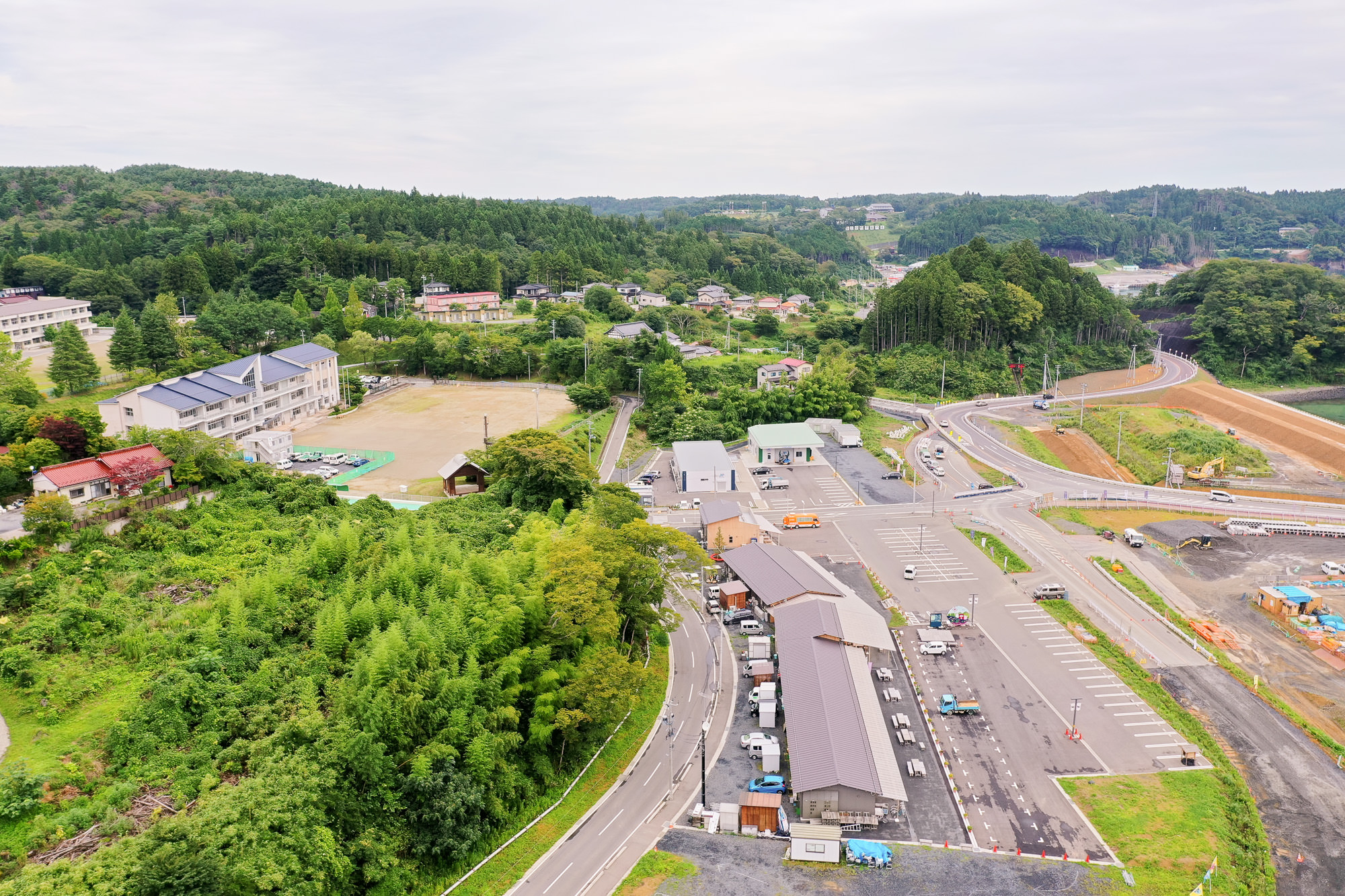 手前に駐車場とプレハブの建物があり、奥に学校とグラウンド、さらに住宅地と森が広がる山間の小さな町の一部を上空から見た写真