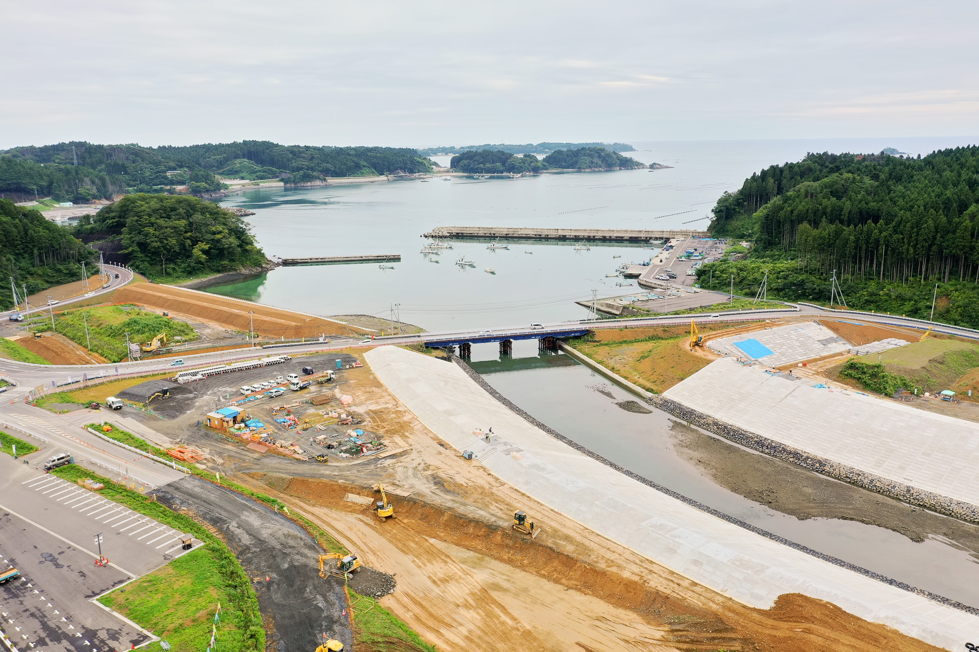 湾に面した漁港と防波堤のある海が見え、手前に川と橋、その周囲で重機が工事を進めている堤防建設中の写真