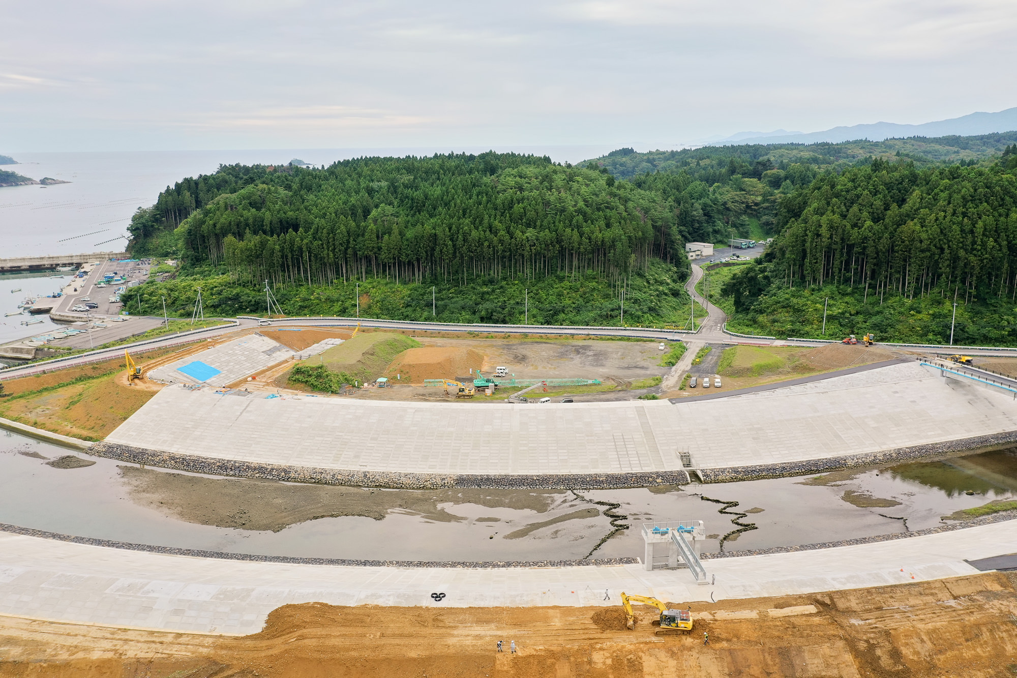 コンクリートで固められた大きな堤防が湾を取り囲み、奥には山と細い道路、手前には重機が作業をしている様子が見える工事現場の写真