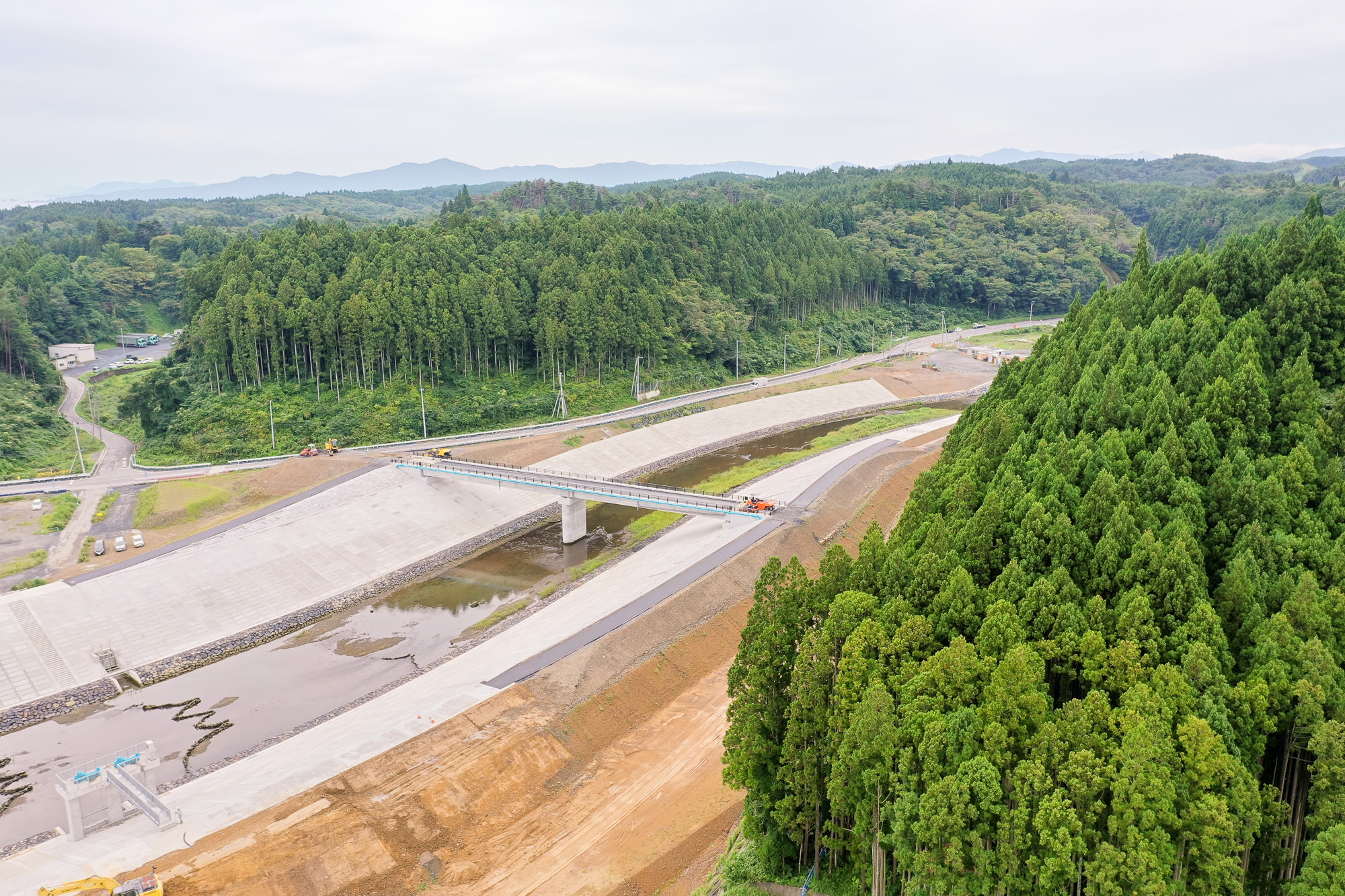 緑の山々に囲まれた谷間を川が流れ、両岸にコンクリートの堤防と新しい橋がかかっている様子を斜め上から見た写真
