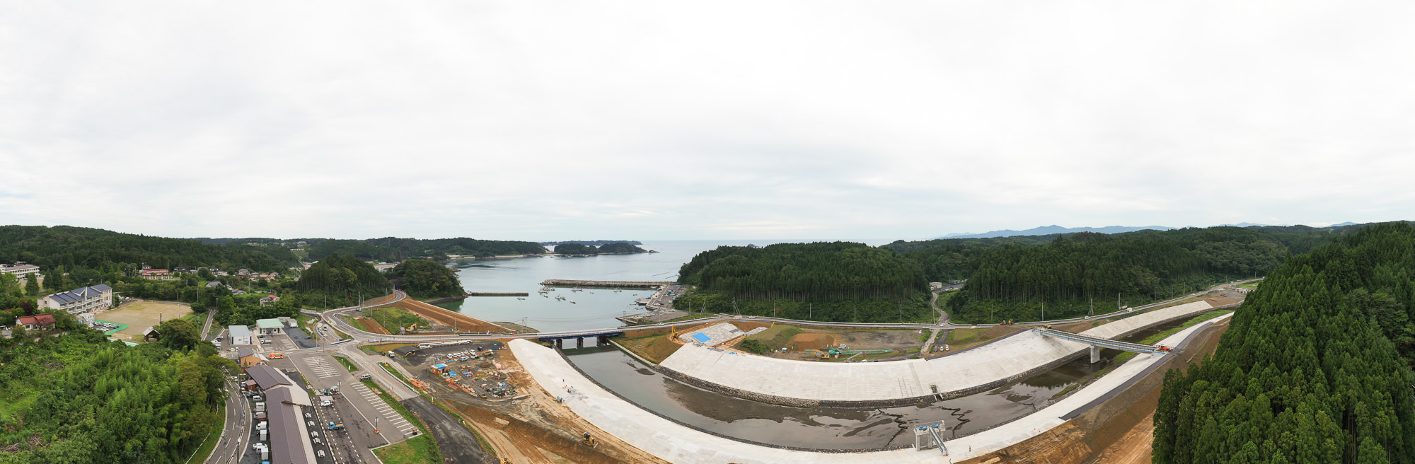 港と山に囲まれた湾の奥に大きな堤防と川が見え、手前には建設中の道路や重機が並ぶ町全体を広く見渡した風景の写真
