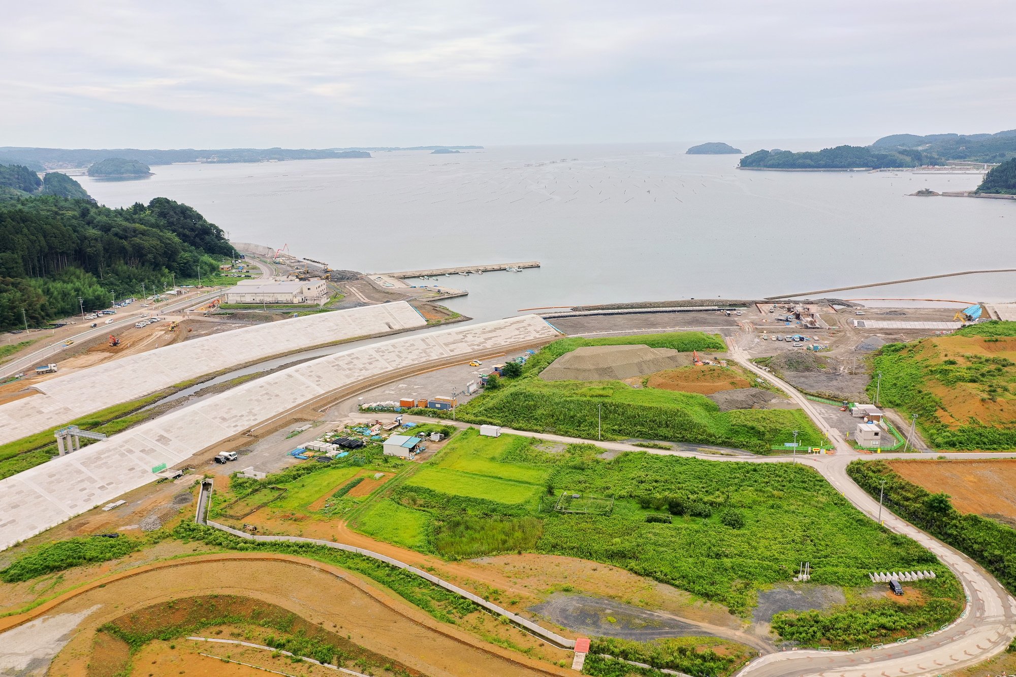 海に面した湾の奥に広がる防潮堤工事現場で、左側に森林が広がり、右側には工事用の重機や土砂が見え、湾内には養殖のための浮きが多数並んでいる様子が写っている防潮堤の建設中の海辺の写真
