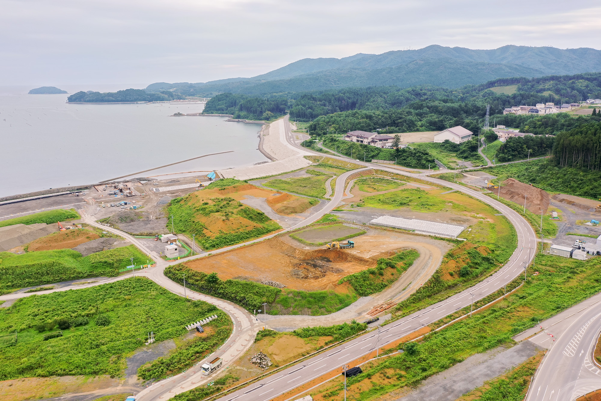 海沿いの防潮堤と湾を右手に見ながら、丘の上の造成地やカーブを描く新しい道路、山間に建つ建物や森が背景に広がる防災インフラ整備中の海岸沿いの写真