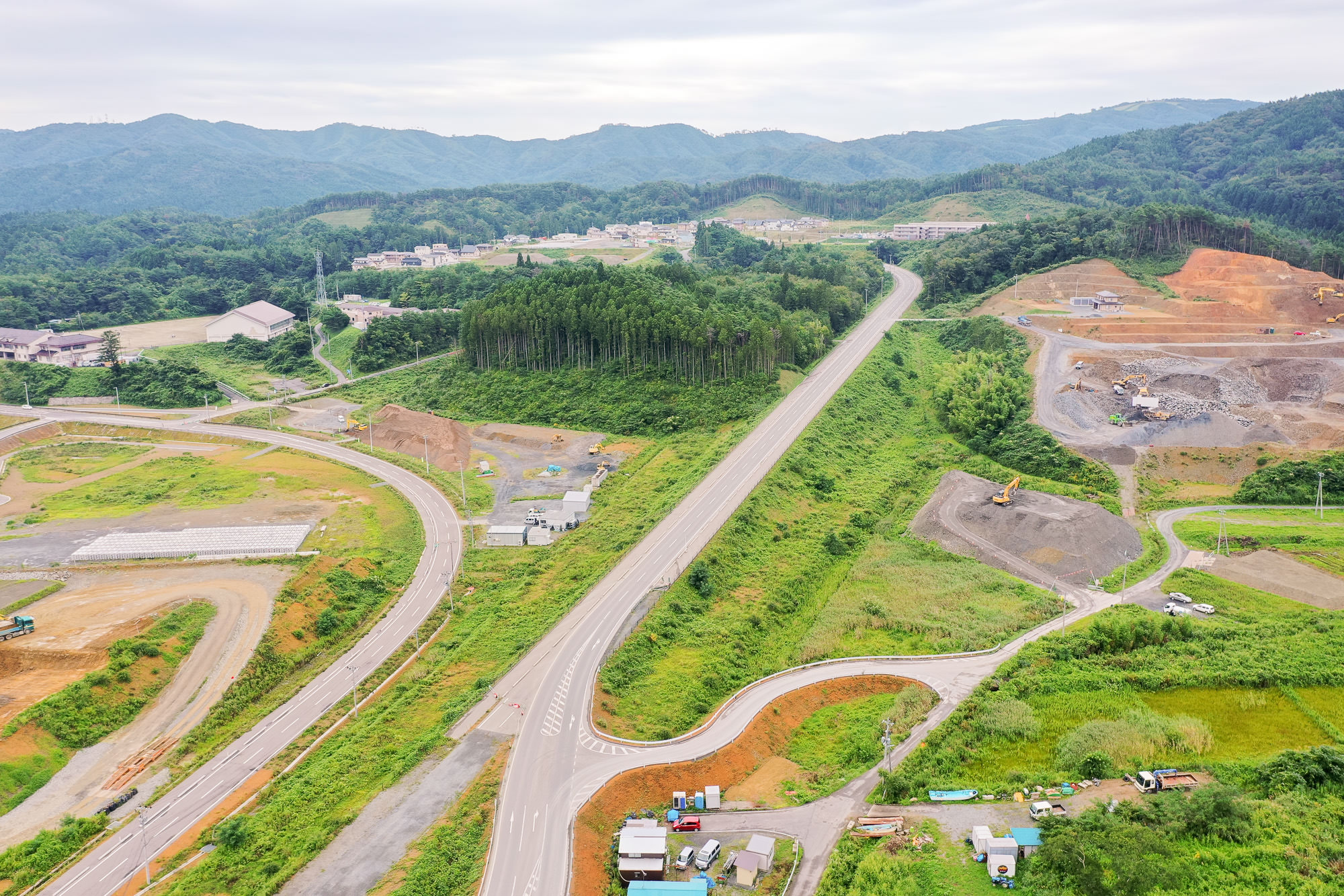 山に囲まれた地域で、新しく整備された広い道路が奥へ続き、両側に造成中の土地や掘削中の現場、整地された区画や森が見える復興工事が進む山あいの町の写真