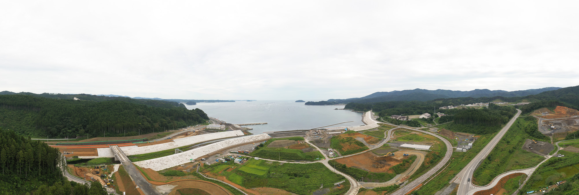 前景に舗装道路と工事用重機、左側に緑深い森林、背景に海岸と小島が広がる空撮写真